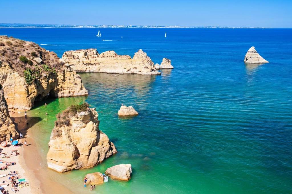 Scenic coastal view with rocky cliffs and sailboats on a clear blue ocean.