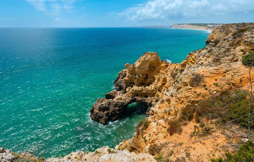 Cliffside view of ocean with rocky formations and blue sky.