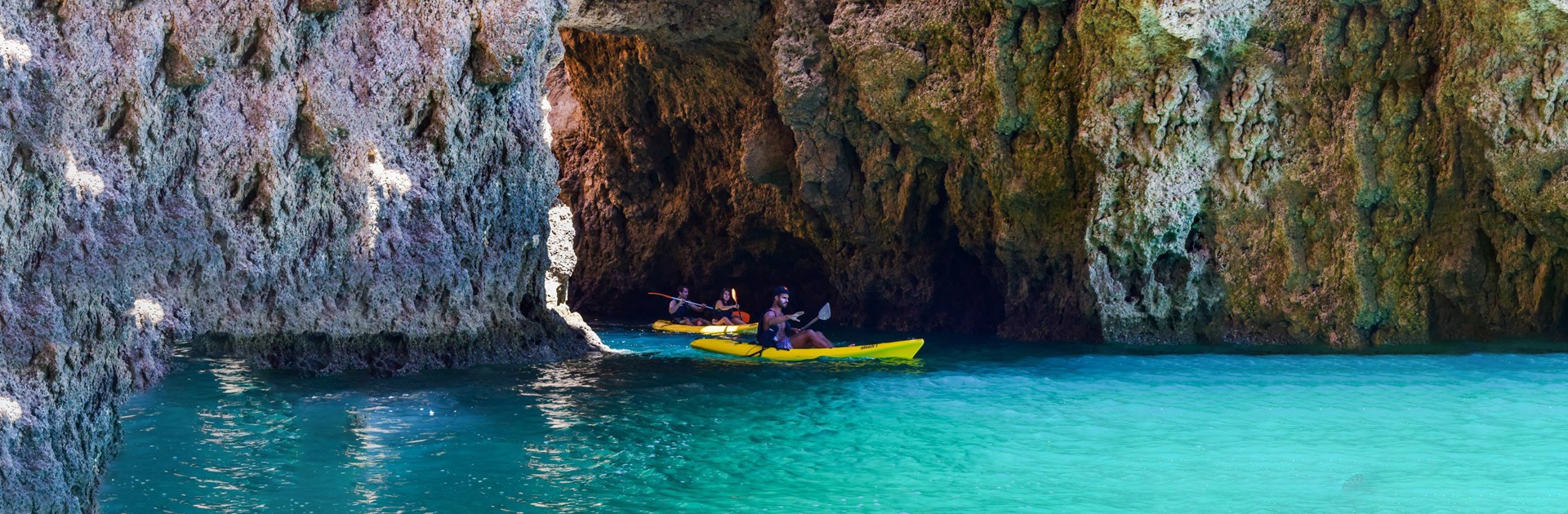 Three kayaks entering a rocky cave by clear turquoise water.