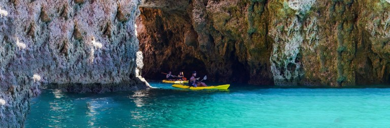 Three kayaks entering a rocky cave by clear turquoise water.